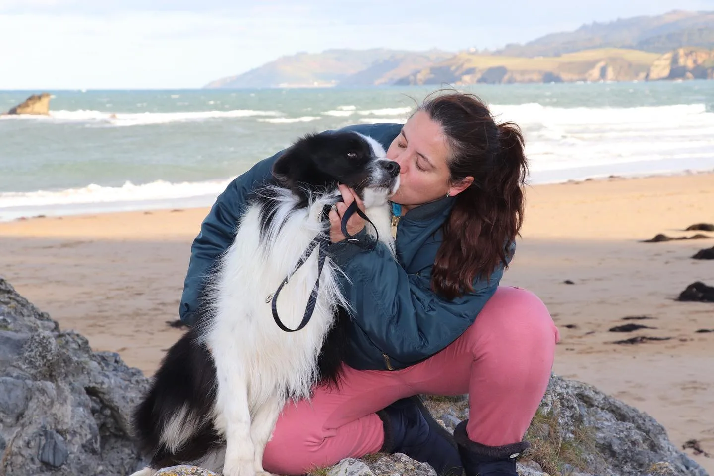 se muestra un border collie de color blanco y negro y a su dueña dándole cariños, están encima de una roca y de fondo se puede ver arena y el mar, están en la playa